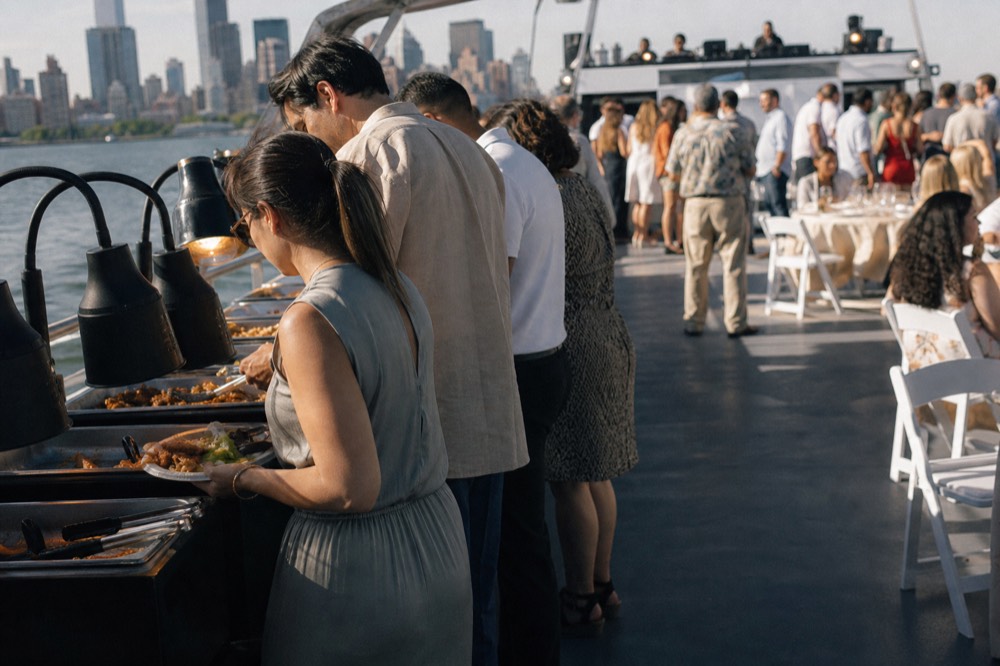 Guests filling plates along a lit buffet counter on a moving boat, dance floor and DJ booth blurred behind them