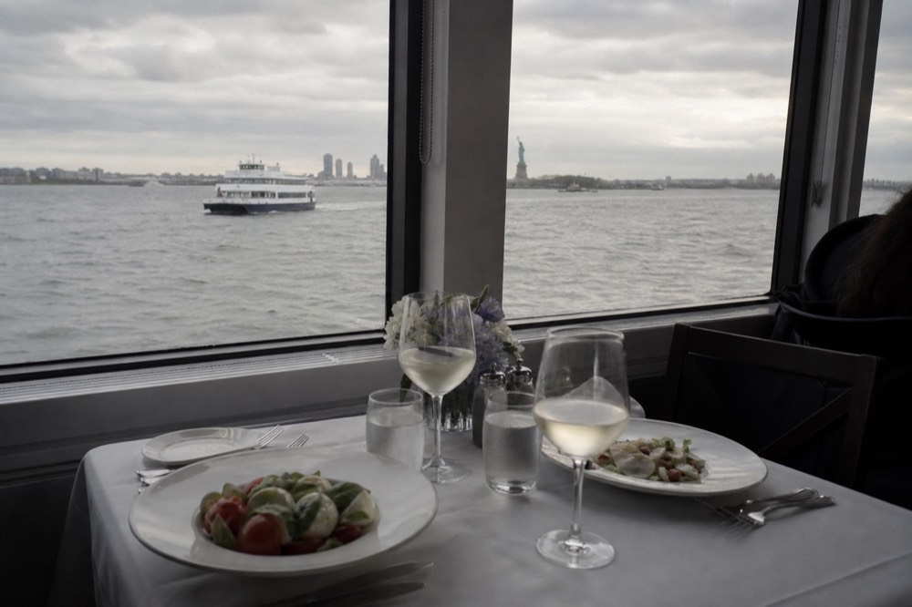 A plated first course set on white linen at a window table aboard a boat, a second setting opposite, dark water outside