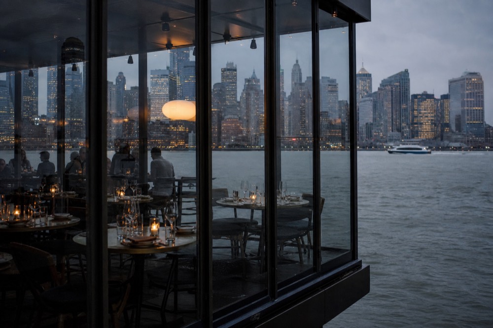 A glass-walled dining deck at blue hour, tables set with candles and water glasses, skyscrapers reflected in the windows.