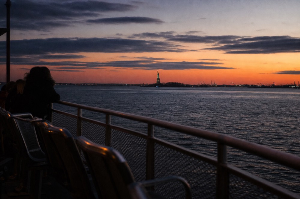 An open upper deck at golden hour with empty railing seats, the harbor water bright and the Statue of Liberty small in the distance.