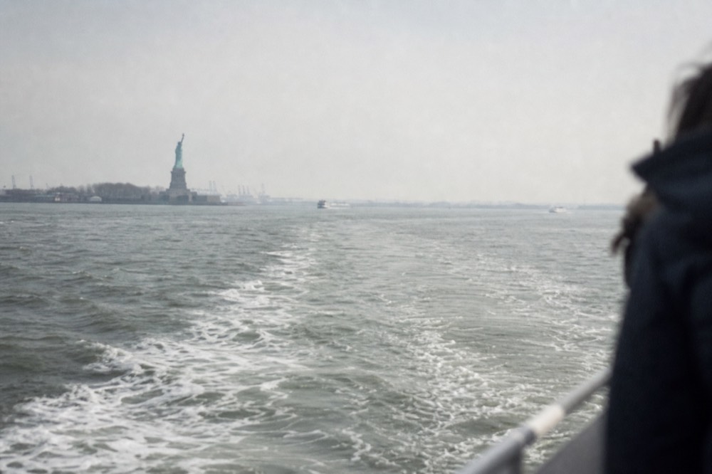 A distant view of the Statue of Liberty from a moving boat rail, small against grey harbor water, a wake curving away in the foreground.