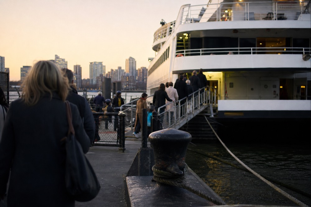 A cruise vessel gangway at a Hudson River pier at dusk, guests boarding in coats, mooring lines and bollards in the foreground