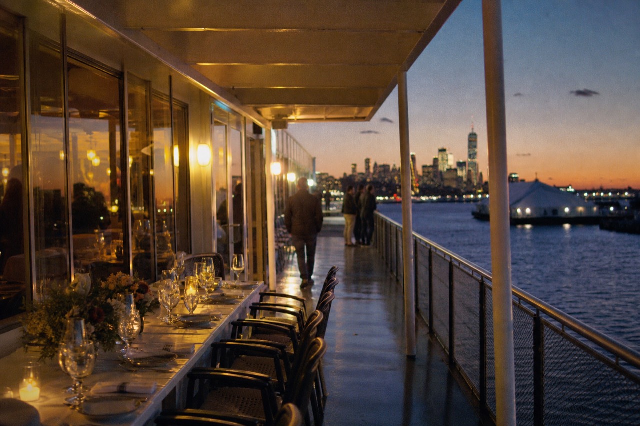 A long table set for a group on a cruise vessel at dusk, wine glasses and place settings, harbor lights beyond the windows