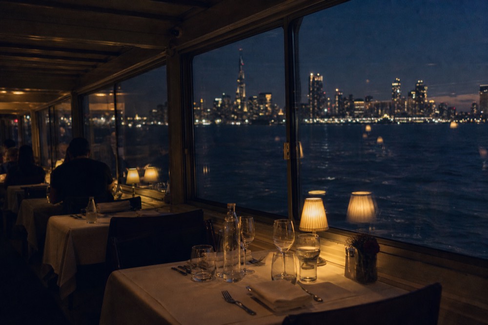 Interior of a dining vessel at night showing separated tables with linen and low lamps, each set apart from the next along a window wall