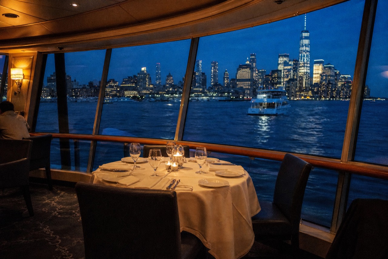 A set table for four beside a curved glass wall on a harbor vessel, empty chairs turned outward toward the lit city skyline at night