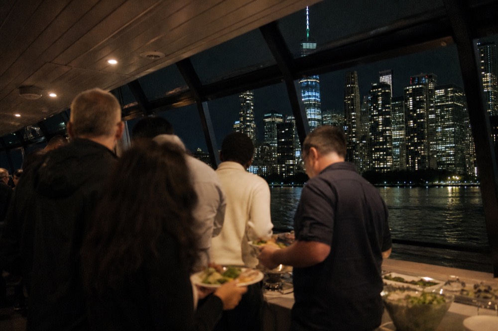 A buffet line on a boat with guests in casual clothes holding plates, warm interior lighting and a dark harbor visible through the windows.