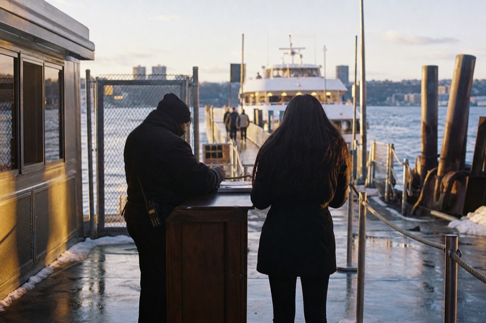 A ticket check point at the head of a boarding ramp in the evening, rope stanchions and a lit vessel waiting at the end of the pier.