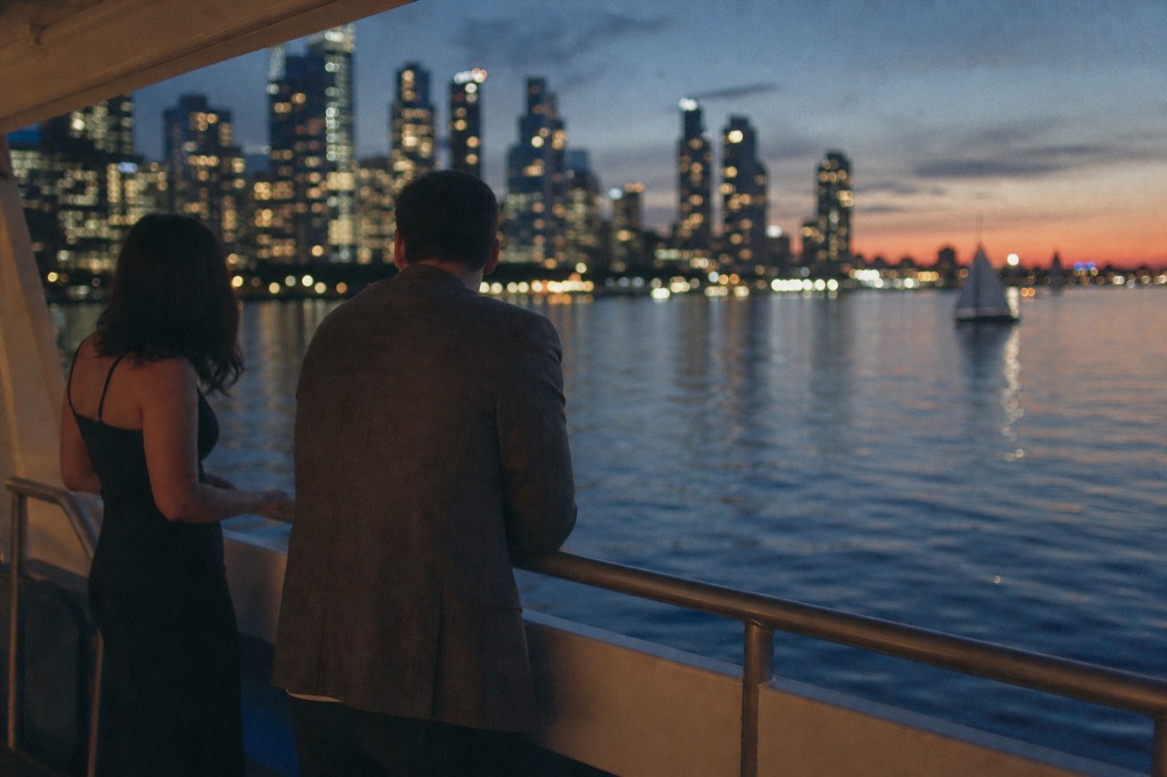 A woman in a dark cocktail dress and a man in a jacket standing at a boat rail at dusk, city lights blurred across the water behind them.