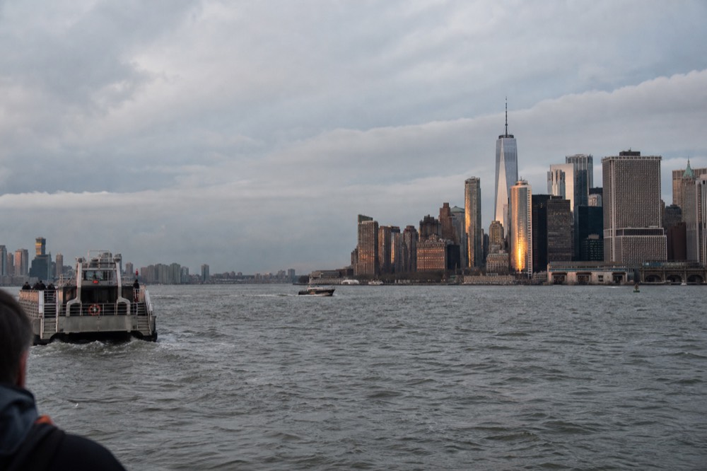 Late golden light striking the glass faces of Lower Manhattan towers, viewed across choppy harbor water from the west