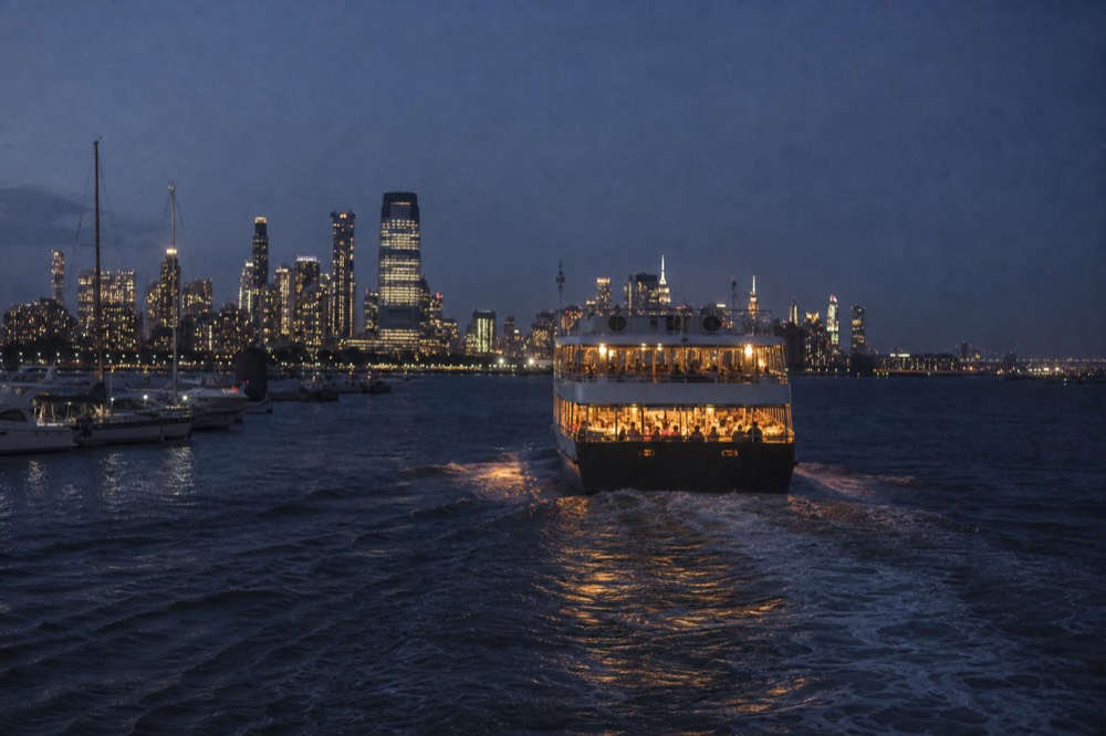 A dining vessel lit from within pulling away from a marina dock at blue hour, wake spreading toward the camera