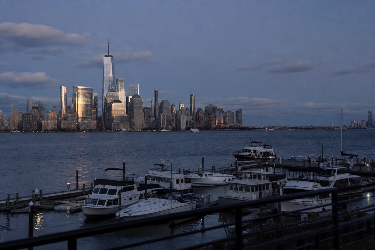 The full Manhattan skyline seen head-on across open water from a marina on the New Jersey shore, towers catching low evening light