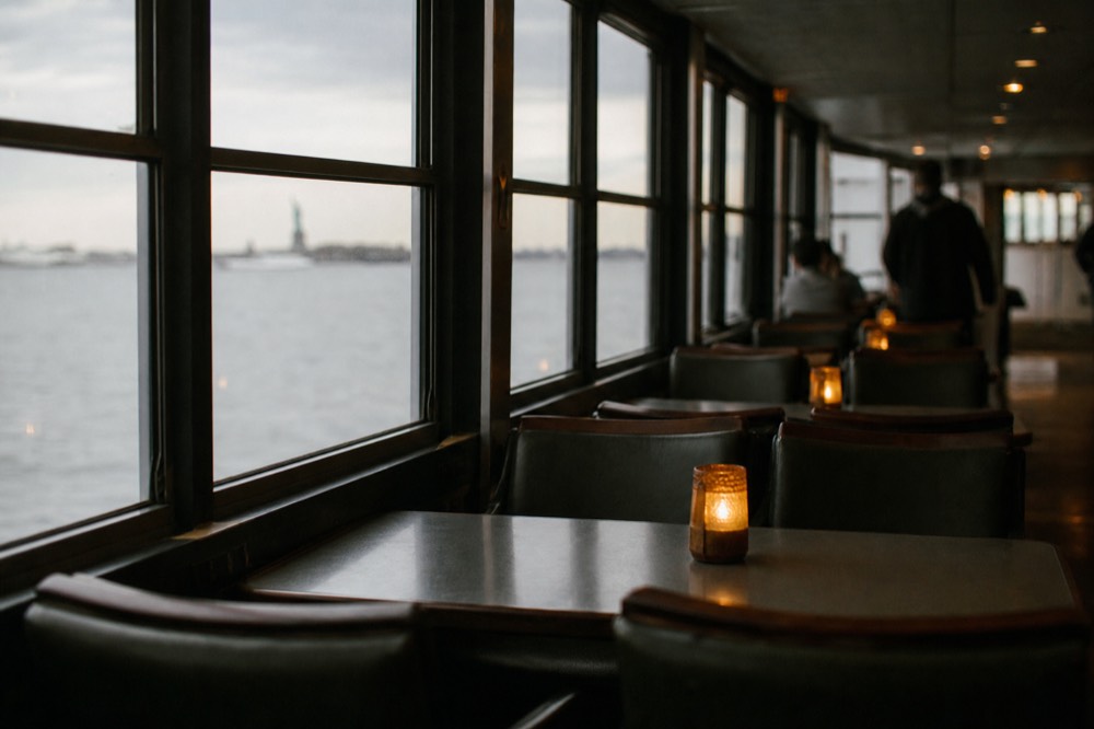 Empty window seats along the glass side of a dining boat, harbor water and a distant shoreline visible through them
