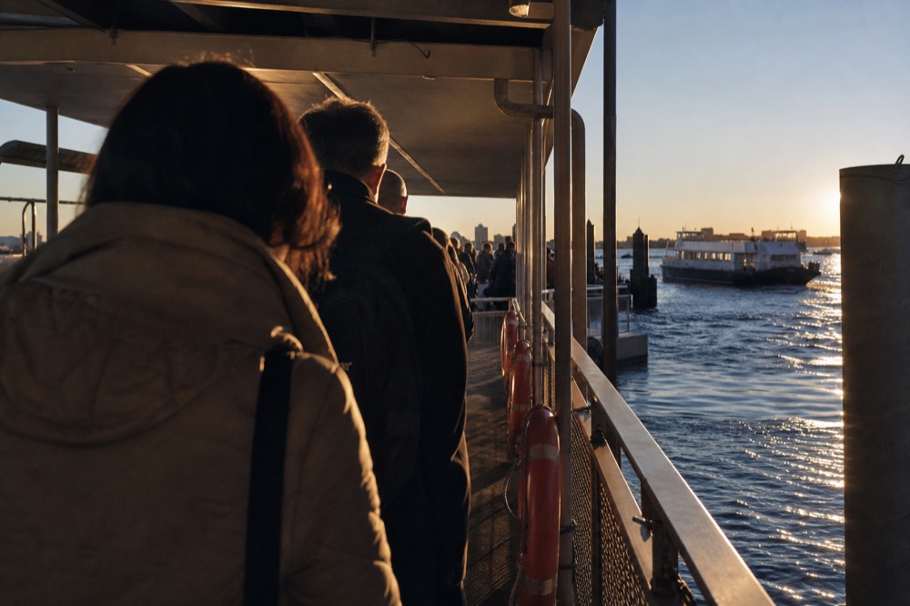 Passengers queueing along a covered pier walkway toward a boarding ramp in early evening light, life rings mounted on the rail.