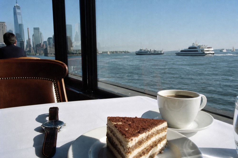 A wristwatch resting beside a dessert plate and a coffee cup on a white tablecloth, dark harbor water and distant lights through the window.