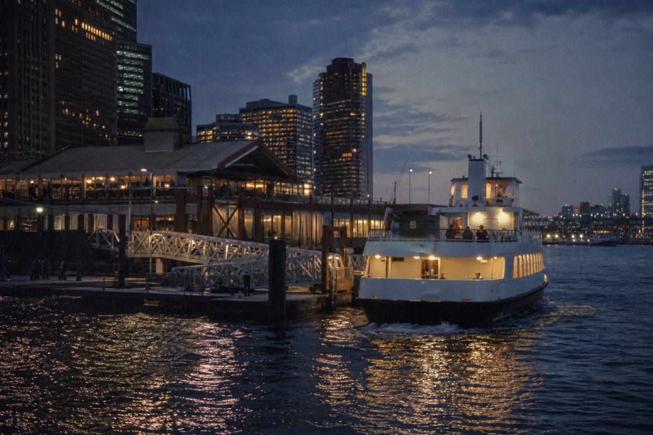A lit harbor boat pulling away from a floating dock at dusk, gangway still visible, city towers rising behind the pier sheds.