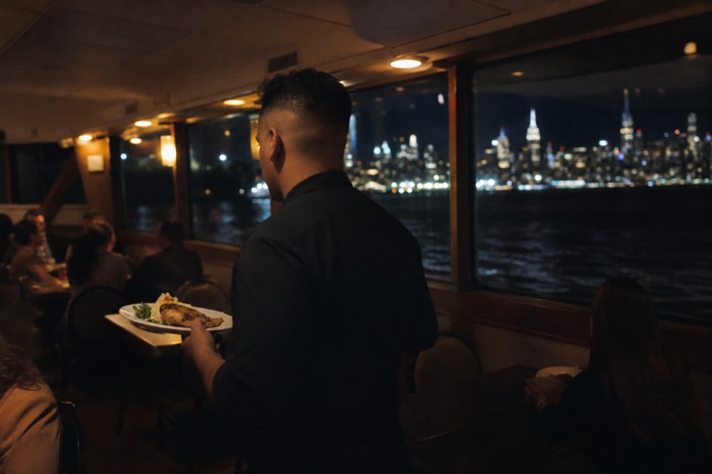 A server carrying two plated dinners between tables on a moving boat, warm interior lighting, dark windows behind