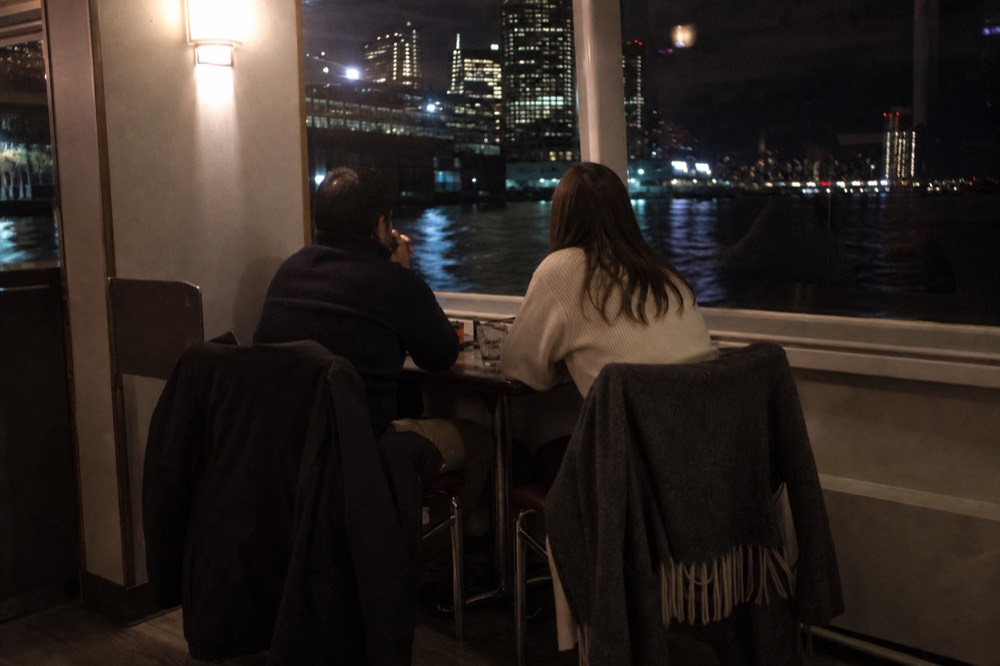 Two people at a small table by a boat window at night, coats over the chair backs, city lights blurred behind the glass
