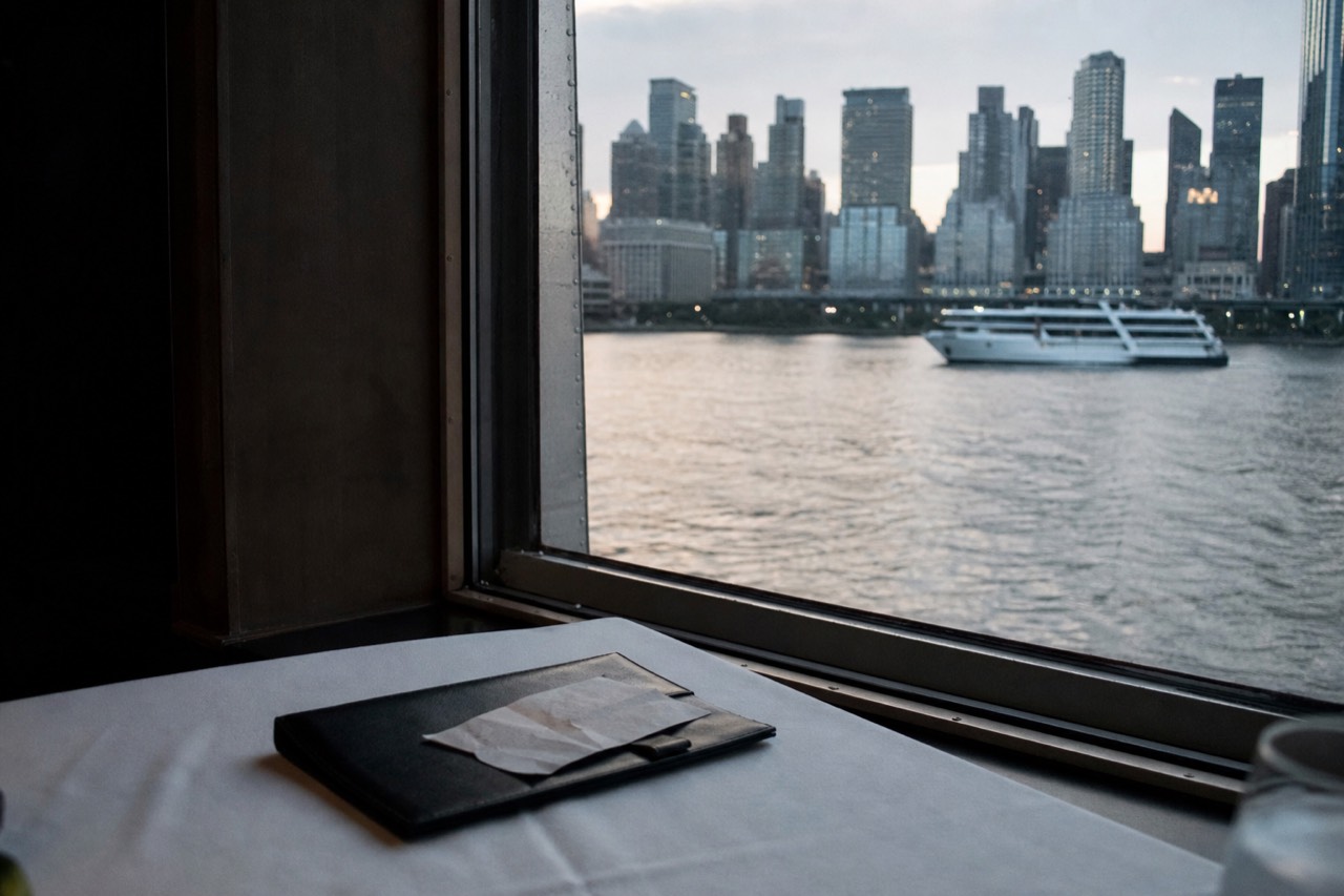 A leather check folder and a folded receipt on a white tablecloth by a ship window, water and evening city light outside