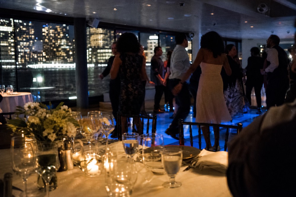 Passengers dancing on a lit dance floor inside a harbor vessel, city lights blurred through the window wall behind them