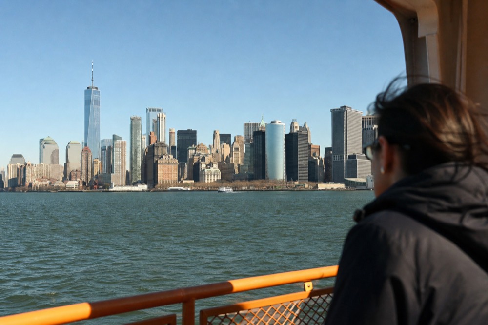Midday view from a boat deck of the Manhattan skyline in clear daylight, individual towers sharply defined above flat green water