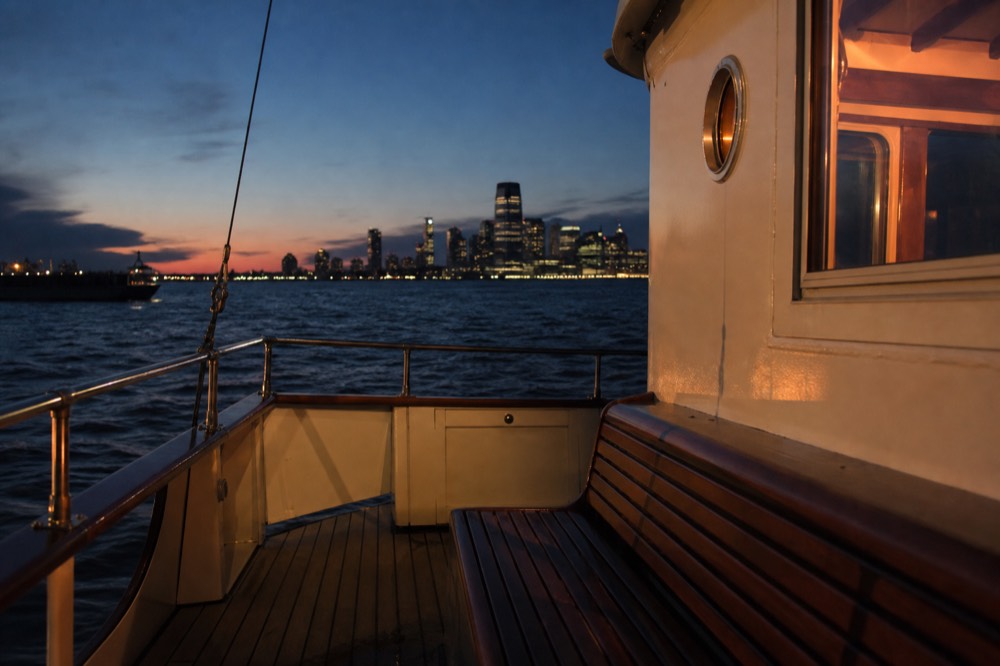 The open teak deck of a classic motor yacht at sunset, empty varnished bench, low orange light across dark harbor water