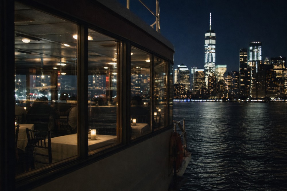 A glass-walled dining saloon on a harbor vessel at night, tables set with white linen and single candles, lit towers filling the windows