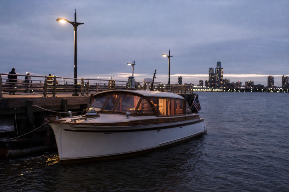 A small classic motor yacht moored at a Hudson River pier at blue hour, warm cabin light through its windows, calm water in the foreground