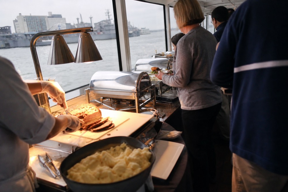 A buffet service line on a cruise vessel with carving station and chafing dishes, guests plating food, harbor water visible through windows behind