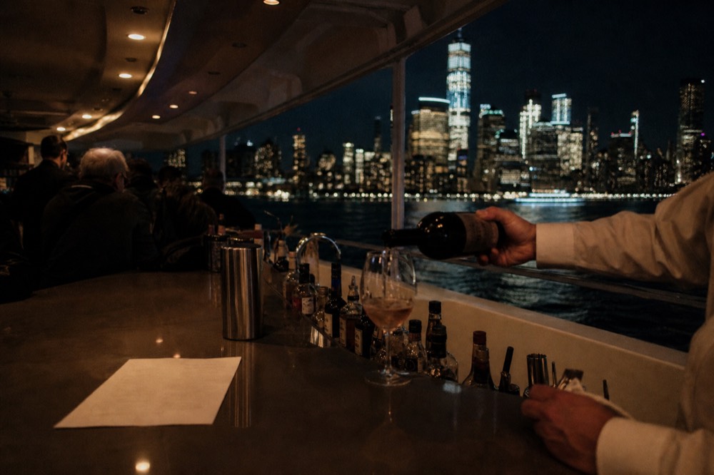 A bar counter on a cruise vessel with a bartender pouring wine, a printed drinks list face down, dark water and distant lights through the window