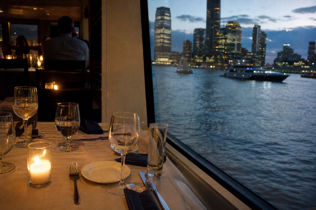 A set dinner table beside a tall window on a harbor cruise vessel at dusk, city lights softened by the glass