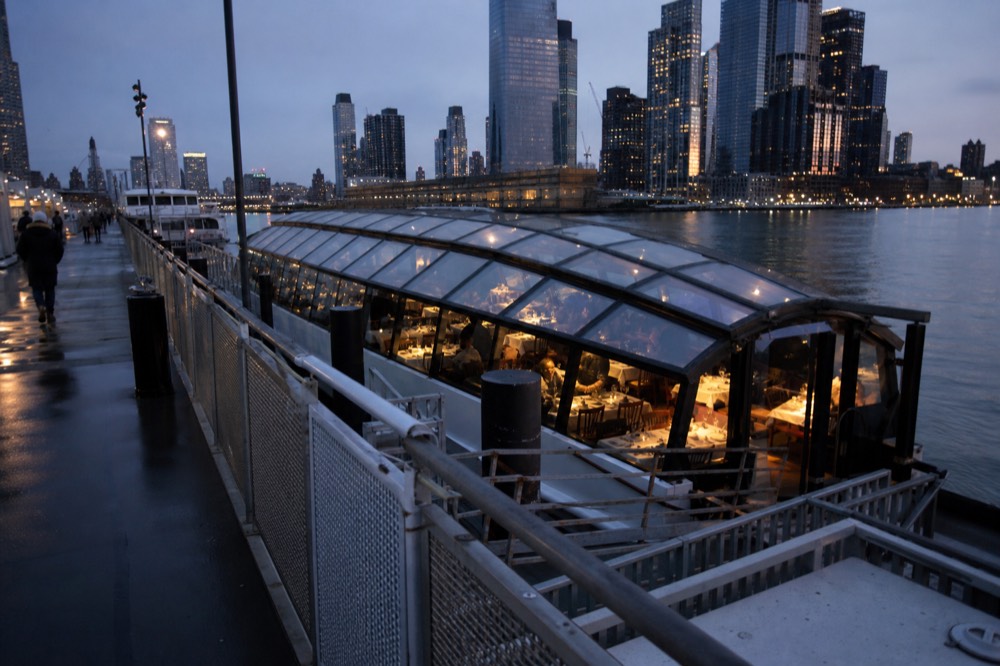 An all-glass dining vessel seen from the pier at blue hour, warm interior light showing set tables through the curved glass roof