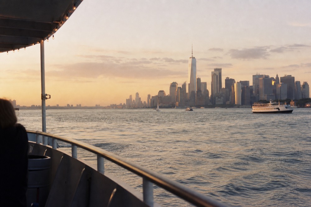 Open upper deck of a small harbor yacht at sunset, empty railing in the foreground and the Lower Manhattan skyline in soft orange haze