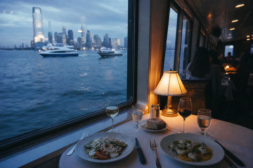 Two place settings side by side on a linen tablecloth aboard a glass-walled boat, one lit by daylight through the window and one by evening lamplight