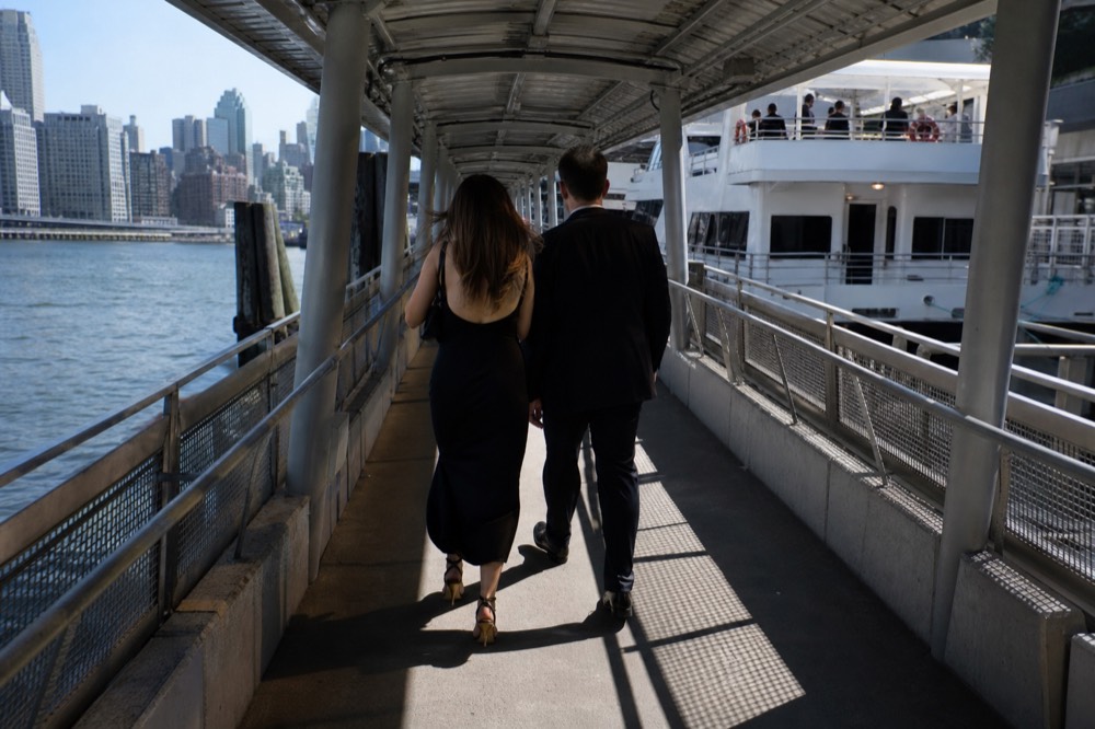 Couple in semi-formal evening dress walking along a covered pier gangway toward a lit boat, seen from behind