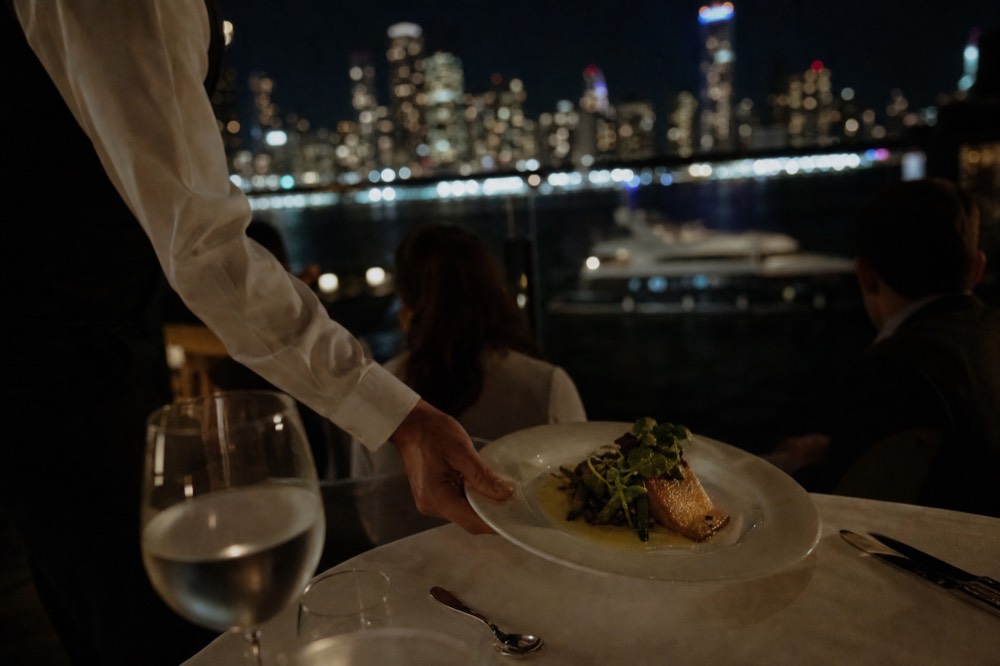Plated main course being set down on a white tablecloth by a server in a waistcoat, blurred harbor lights through glass behind