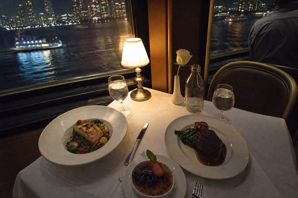 Overhead view of a plated three-course dinner on a white tablecloth beside a cruise vessel window at night