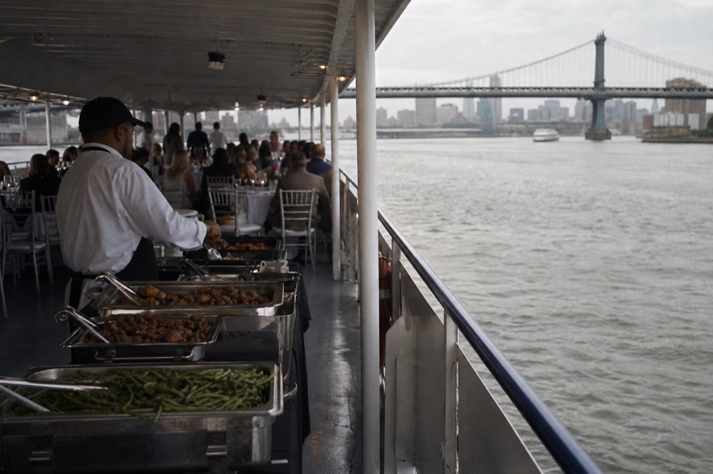 A buffet line with chafing dishes and a server on the lower deck of a dinner cruise boat, guests seated at round tables in the background
