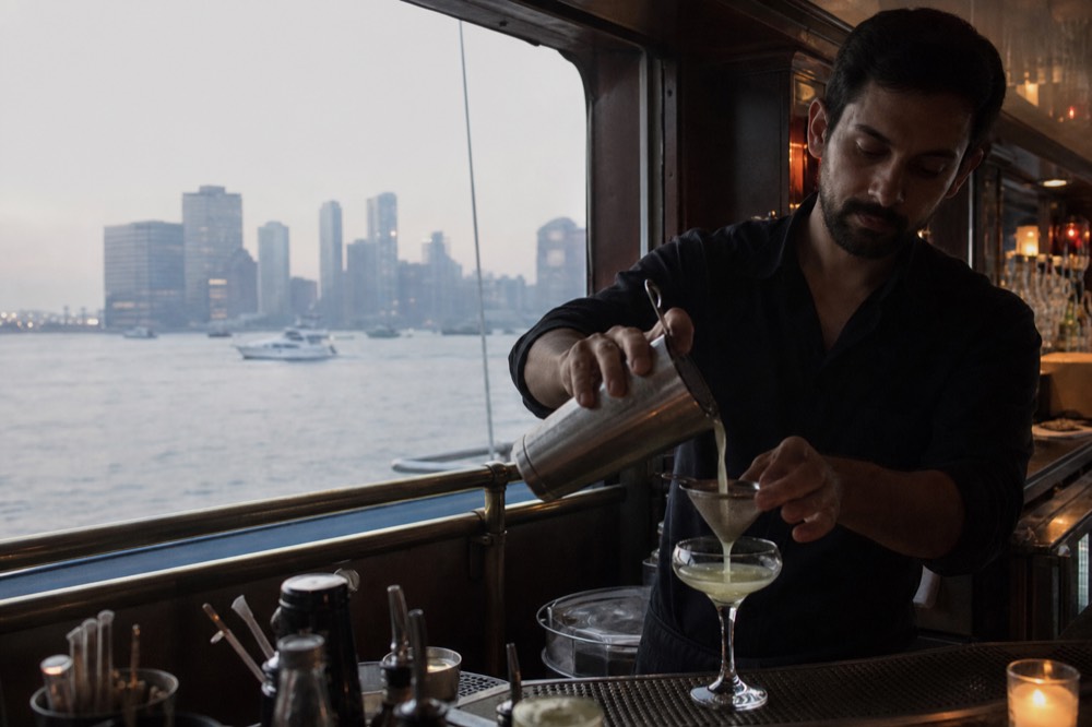 A bartender pouring a cocktail at a small onboard bar with brass rails, harbor lights visible through the window behind