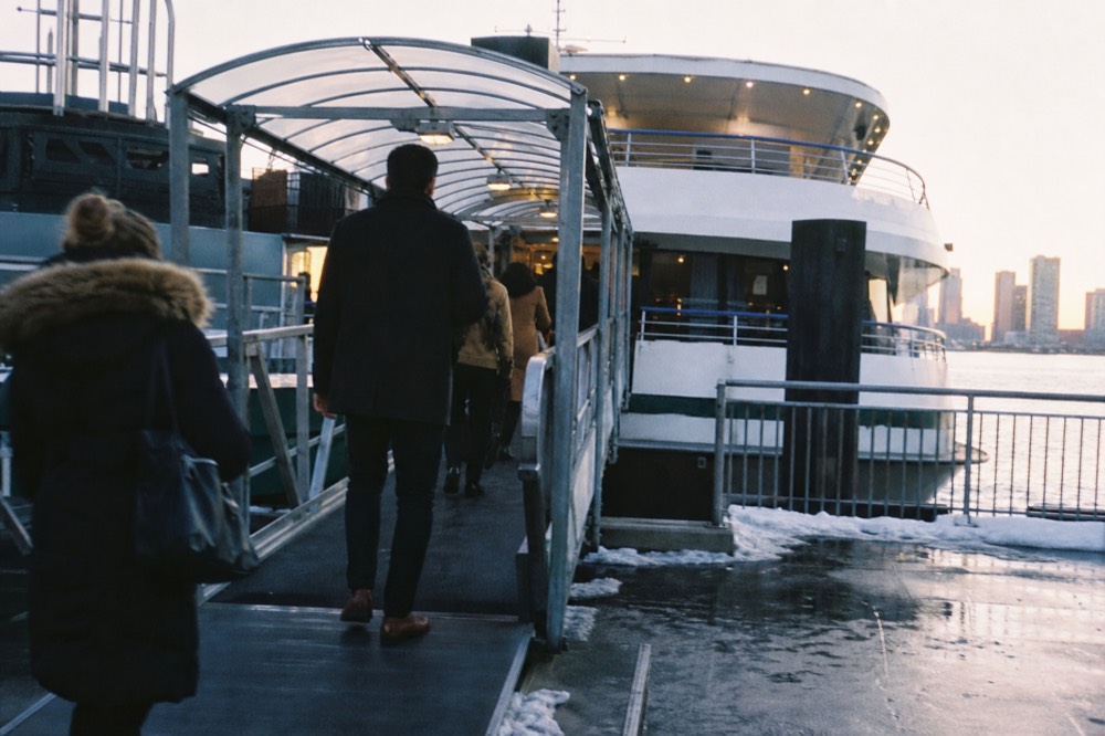 Passengers walking up a covered gangway onto a large white dinner cruise vessel at a Manhattan pier in early evening light