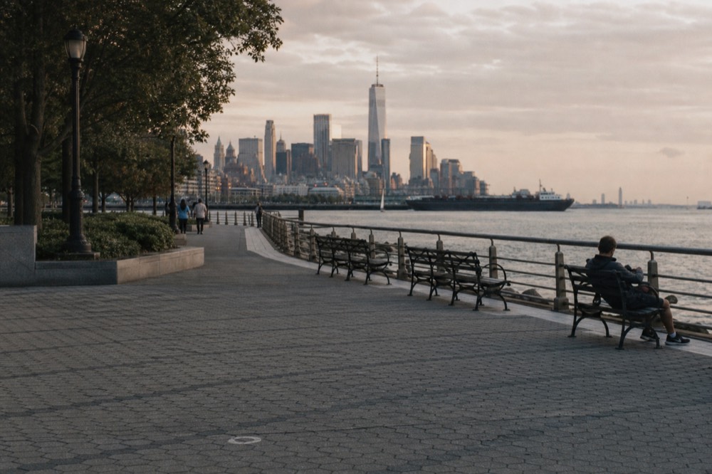 Wide waterfront promenade in a harbourside park with benches facing the water and the Manhattan skyline on the horizon