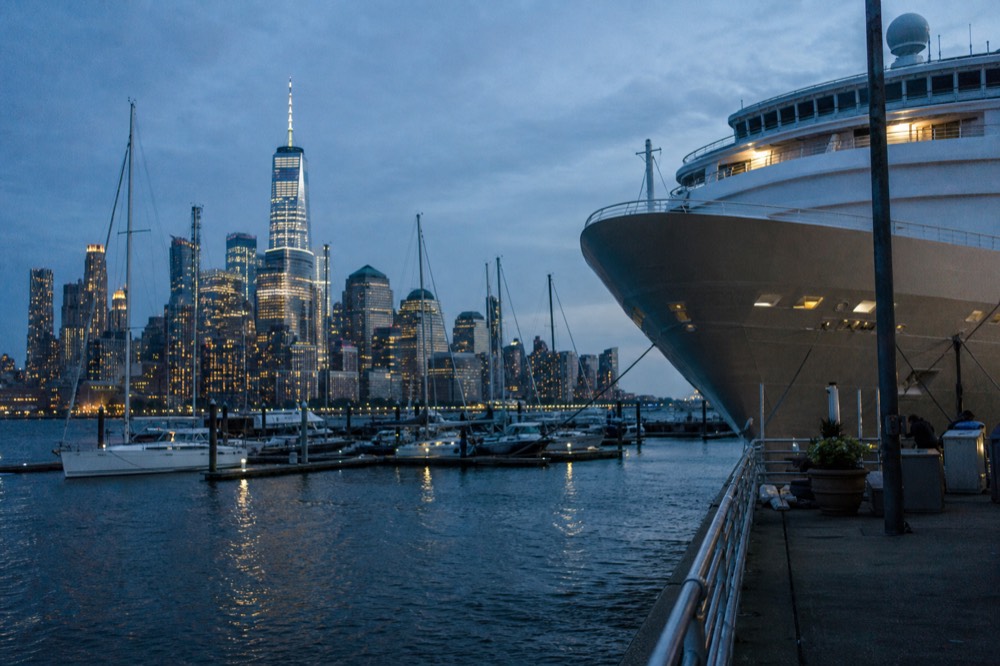 Large white cruise vessel moored at a marina dock with sailing yachts alongside and the Manhattan skyline across the water behind