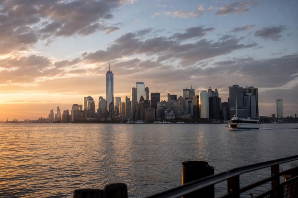 Wide unbroken view of the lower Manhattan skyline across calm harbor water in evening light, entire skyline visible in one frame