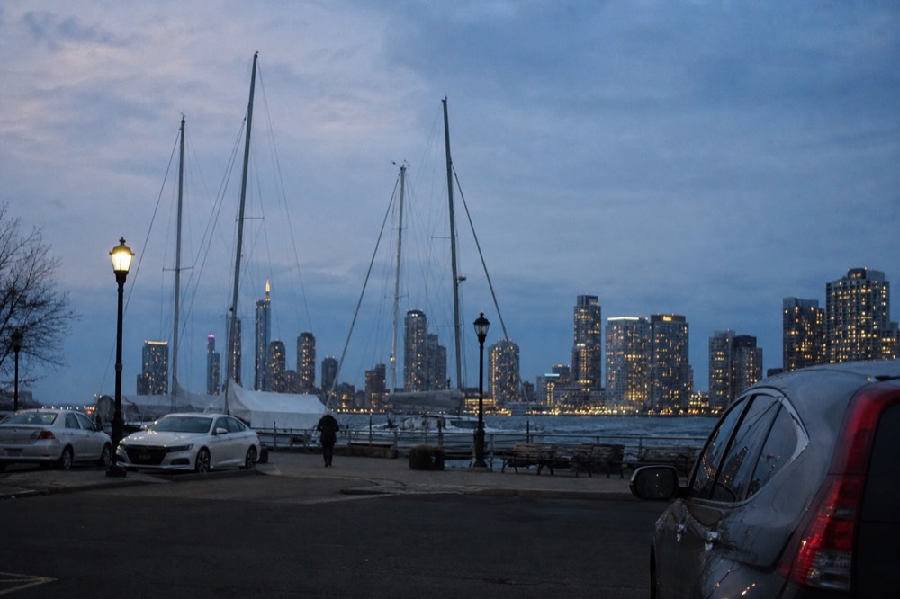 Marina parking area beside a waterfront walkway with a few cars and boat masts, city towers across the water in the distance