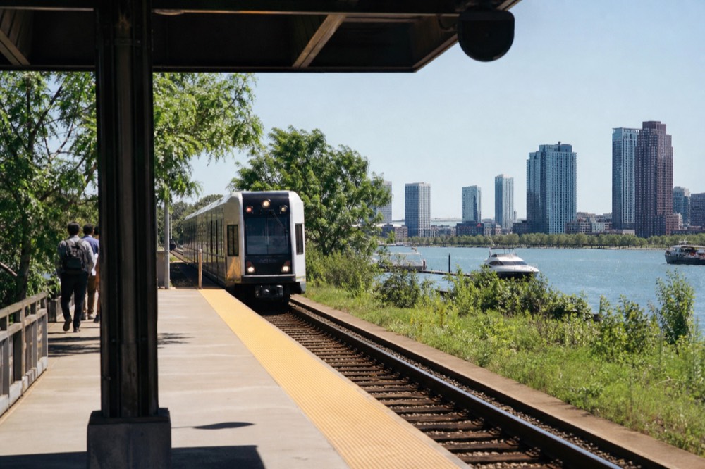 Light rail train arriving at an open-air station platform in a park setting with waterfront trees and distant city towers