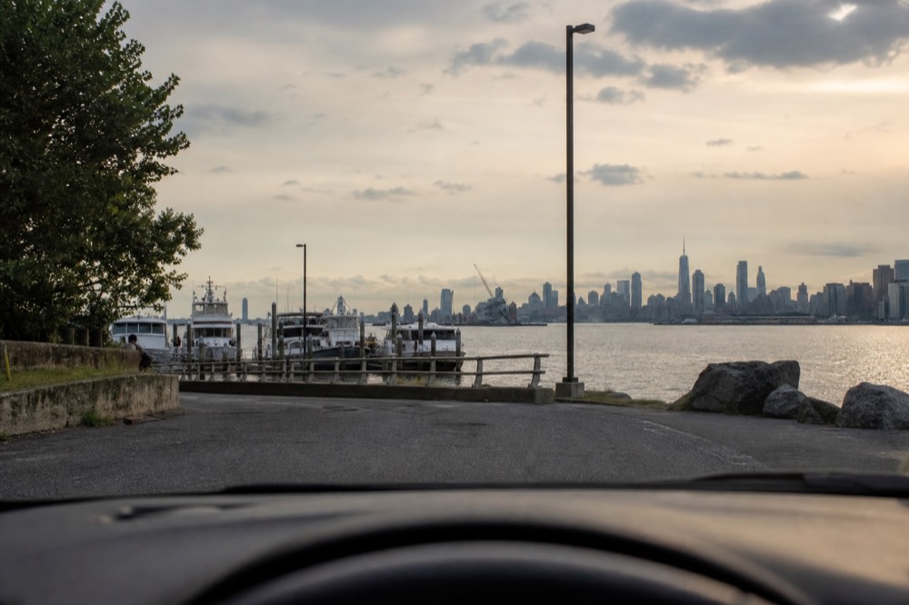 Car pulling into a quiet waterfront parking area at golden hour with a marina and distant skyline visible ahead