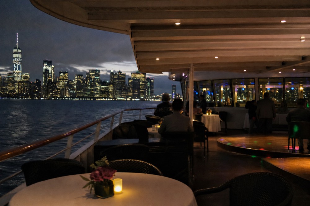 Buffet dining deck on a cruise vessel with round tables, a small dance floor and large windows showing water at dusk