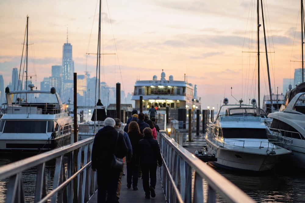 Passengers walking along a floating marina dock toward a lit cruise vessel at dusk, boat masts either side