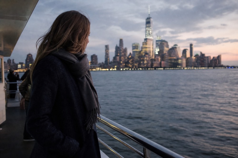 Person in a wool coat and scarf standing at a yacht rail in fading winter light, city lights beginning to show behind