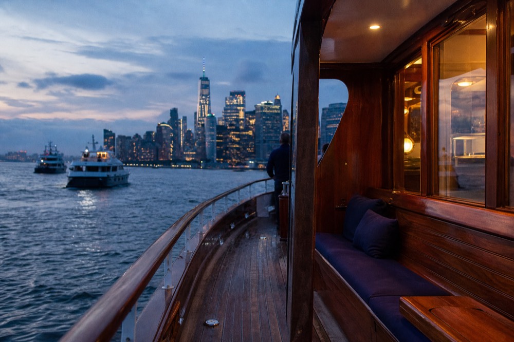 Varnished teak afterdeck of a classic motor yacht with wooden bench seating and a glass-walled saloon behind, harbor water beyond