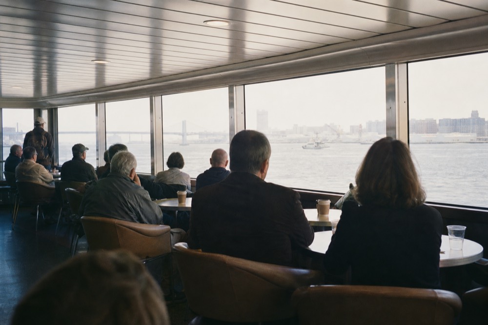 Guests seated at round tables on the enclosed lower deck of a large cruise vessel in daylight, harbor water and a distant bridge through the windows